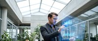 Professional businessman analyzing holographic financial data and charts in modern office environment with natural lighting and glass architecture.