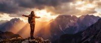 Young woman hiker with backpack standing on mountain peak with arms outstretched celebrating summit achievement during golden hour sunrise in alpine landscape.