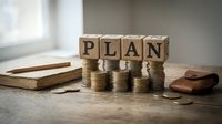 Wooden blocks spelling PLAN with stacked coins and books on desk representing financial planning and business strategy concepts.