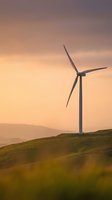 Wind turbine standing on rolling green hillside at golden hour sunset, generating renewable energy against warm sky. Sustainable power generation.