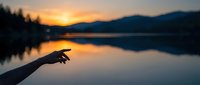 Human hand silhouette pointing towards golden sunset over calm mountain lake with forest reflection creating peaceful outdoor scene for meditation concepts.