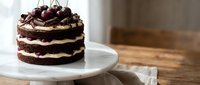 Decadent chocolate layer cake with cream frosting topped with fresh cherries on white plate, rustic wooden background for bakery dessert menu designs.
