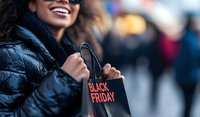 Young African American woman smiling while holding shopping bag during seasonal sale, wearing winter puffer jacket. Selective focus on shopping bag with blurred background.