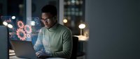 Young African American man working late at night on laptop with digital gear icons and technology interface elements in modern office environment.
