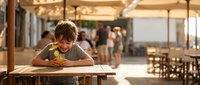 Young boy enjoying meal at outdoor beachside restaurant during golden hour with warm ambient lighting and blurred diners in background.