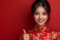 Young Asian woman wearing traditional red and gold qipao dress with floral pattern showing thumbs up gesture against red background, bright smile and festive makeup.
