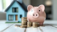 Pink ceramic piggy bank with stacked gold coins in front of blue house model on wooden surface, representing savings and home investment goals.