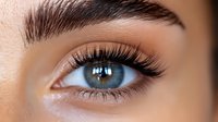 Close up macro shot of feminine eye with long black eyelashes, defined eyebrow and bright blue iris showing intricate details and textures of human eye.