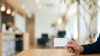 A person with medium skin tone wearing dark clothing holds a blank white business card at a wooden desk in a modern office environment with blurred background and natural lighting.