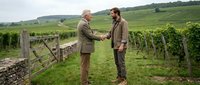 Two middle-aged Caucasian men in countryside attire shake hands near vineyard fence, discussing wine business partnership in rural agricultural setting.