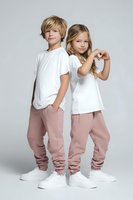 Caucasian boy and girl in matching white t-shirts and pink joggers pose confidently against gray background. Girl makes heart gesture while boy stands casually.
