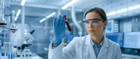 Female scientist in white lab coat and safety glasses holding blood sample vial in modern laboratory setting. Suitable for medical, healthcare, and research applications.