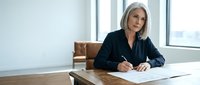 Mature woman with white hair and light skin tone, wearing dark blazer, sitting at wooden desk writing on document in modern office with floor-to-ceiling windows, professional business setting for corporate communications.