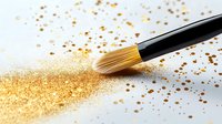 Professional makeup brush sweeping through scattered gold glitter powder on white surface, creating dynamic motion trail for cosmetic and beauty photography.