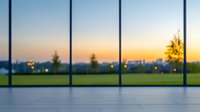 Modern office interior with floor-to-ceiling windows overlooking green landscape at sunset, blurred city lights in background, minimalist architecture design space.