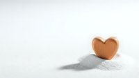 Wooden heart shape sitting on white sand pile against clean white background, minimalist love concept for romantic designs and wellness themes.