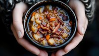 Hearty beef and potato soup in black ceramic bowl held in hands, rich broth with tender meat chunks and vegetables, moody dark food photography.