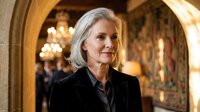 Confident mature businesswoman with silver hair in elegant black suit standing in luxurious hotel lobby with ornate architecture and warm lighting.
