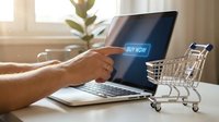 Hand pressing a glowing buy now button on a laptop screen next to a mini shopping cart, symbolizing online shopping and e-commerce.