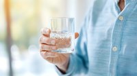 Male hand holding crystal glass with clear water against blurred bright background, wearing light blue casual shirt. Fresh drink concept for healthy lifestyle.