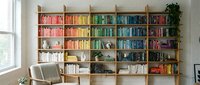 Modern home library with color-coordinated books arranged by rainbow spectrum on wooden shelving unit, bright reading nook with comfortable chair and plants.