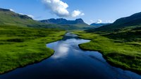 Meandering river flows through vibrant green valley with dramatic mountain peaks and blue sky. Pristine wilderness landscape showcases untouched nature and serene environment.