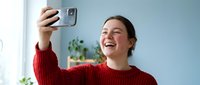 Happy young woman taking selfie with smartphone at home wearing red sweater, smiling and laughing while using mobile device for social media content creation.