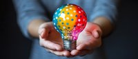 Colorful light bulb painted in rainbow colors with polka dot pattern held gently in cupped hands against dark background, representing creativity and innovation.