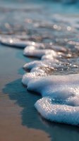 Gentle ocean waves creating white foam patterns on sandy beach with sparkling bokeh water reflections in soft morning light, macro close-up view.