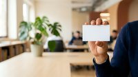 Mature male professional in dark blue sweater holding blank white business card in modern office environment with green plants and colleagues working at desks in background for corporate branding.