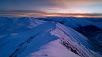 Majestic snow-covered mountain peaks at sunset with dramatic sky casting purple and orange hues across pristine winter landscape in remote wilderness.
