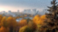 Raindrops on window glass with blurred autumn forest and city skyline in background creating dreamy bokeh effect during fall season at sunset.