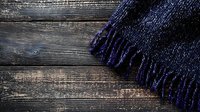 Dark blue wool scarf with fringe lying on rustic wooden planks background, creating contrast between soft textile and rough wood grain texture.