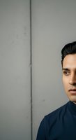 Young man with dark hair and olive complexion wearing navy blue shirt, looking at camera against neutral gray wall with vertical line, suitable for corporate portraits and professional headshots.
