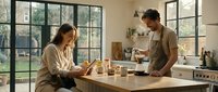 Young couple enjoying morning coffee together in modern kitchen with large windows and natural light creating warm domestic atmosphere.