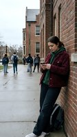 Young Caucasian female college student using smartphone while leaning against brick university building wall with backpack and other students walking in background.