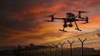 Professional drone flying over security fence at sunset with dramatic orange sky for surveillance and monitoring operations.