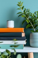 Stack of colorful books on white table with green houseplant in ceramic pot against turquoise wall, creating modern minimalist home office or reading corner setup.