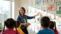 Smiling female teacher with curly hair presenting children's artwork to diverse elementary students in bright classroom with colorful drawings on wall.