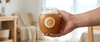 Hand holding glass jar of golden honey with swirl pattern in bright kitchen interior for natural food and healthy lifestyle concepts.