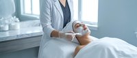 Professional cosmetologist applying facial mask treatment to female client in modern spa salon for skincare and beauty wellness therapy.