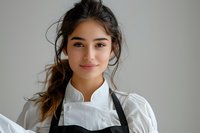 Young Hispanic female chef in white uniform and black apron smiling confidently at camera against light gray background, messy bun hairstyle with loose strands.