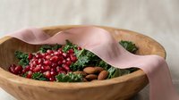 Fresh kale salad with pomegranate seeds and almonds in wooden bowl with pink ribbon for healthy eating and nutrition concepts.