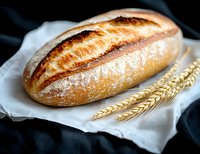 Fresh crusty sourdough bread loaf with golden crust and flour dusting on white linen cloth, decorated with wheat ears against dark background.