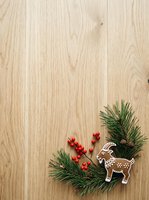 Light wood surface with festive evergreen branches, red berries, and gingerbread reindeer decoration arranged in corner. Overhead flat lay composition ideal for holiday marketing, seasonal greeting cards, and winter promotional materials.