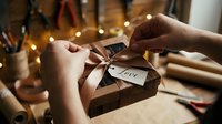 Hands tying a satin ribbon bow on a kraft paper gift box with a love tag, warm bokeh lights in background, cozy handmade wrapping scene.