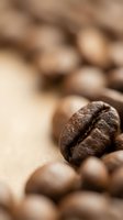 Close-up macro shot of roasted coffee bean with detailed texture and crack, surrounded by blurred beans creating warm brown bokeh background for cafe branding.