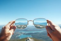 Scenic mountain landscape and lake viewed through metallic frame sunglasses held up against blue sky, creating optical illusion of clear vision perspective.