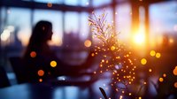 Small decorated Christmas tree with warm golden lights on table in cozy cafe with blurred woman silhouette in background during sunset creating festive atmosphere.