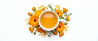 Overhead view of golden herbal tea in white cup surrounded by dried chamomile flowers, petals, and green leaves on white background. Suitable for wellness, natural remedies, and beverage packaging.